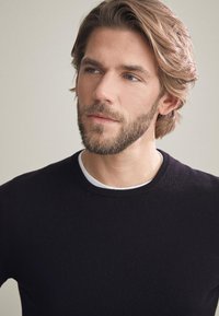 Svart stickad tröja med rund halsringning, lager på lager över en vit innertopp. Mjuk textur och åtsittande design med minimal dekoration.