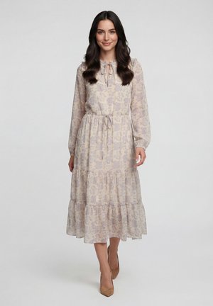 Kvinde med langt mørkt hår, iført en lys blomsterprintet midi-kjole med lange ærmer og bindebånd i nakken samt bælte i taljen, stående foran en ensfarvet baggrund.