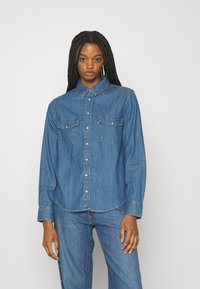 Jeanshemd in mittlerem Blau, mit einem Button-Down-Kragen, zwei Brusttaschen und einem geschwungenen Saum. Weiche Textur mit kontrastierenden Nähten.