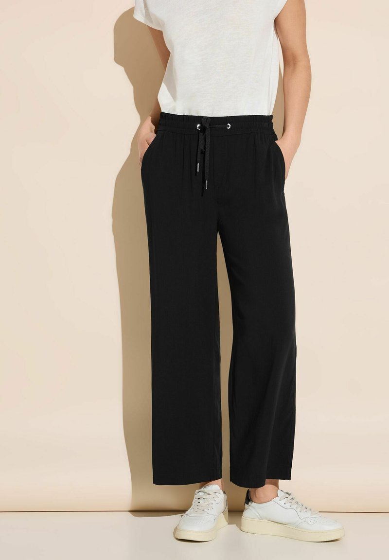 Osoba ubrana w czarne spodnie z szerokimi nogawkami, z elastyczną talią i sznurkiem, w połączeniu z białymi trampkami i białą bluzką z krótkim rękawem.