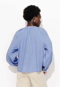 Blouse longue à manches longues bleu clair avec un col froncé et des poignets élastiques. Le tissu texturé offre une apparence douce et fluide.