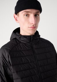 Crna pufasta jakna s horizontalnim kvadratnim uzorkom, visokim ovratnikom i prednjim patentnim zatvaračem. Uparen s crnom pletenom kapom.