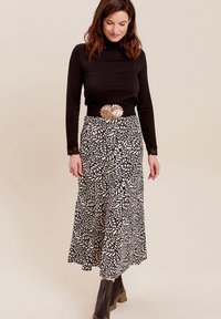 Schwarzer Rollkragenpullover kombiniert mit einem schwarz-weißen Maxikleid mit blätterartigen Mustern. Silberner Gürtel mit floralen Akzenten und braunen Stiefeln.