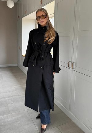 Svart trench coat med dubbelknäppning och knytdetalj i midjan, i kombination med mörkblå jeans och klackskor med spetsig tå, mot en neutral inredning.