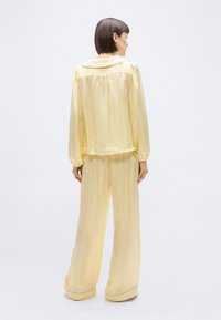 Γυναίκα όρθια με πλάτη, φοράει φαρδιά, ανοιχτό κίτρινο σατέν μπλούζα και παντελόνι με φαρδιά πόδια και γαλάζια διακόσμηση στο γιακά και στο τελείωμα του παντελονι ού.