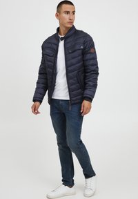 Navy puffer jacket with a stand collar, zip closure, two front pockets, and visible stitching. Paired with blue jeans and white sneakers.