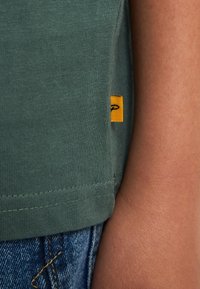 Donkergroen katoenen T-shirt met korte mouwen, voorzien van een geel label met een logo onderaan de zoom. Glad stof, eenvoudig ontwerp.