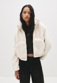 Mlada ženska z dolgimi temnimi lasmi, oblečena v kremno puhasto zadrgo s kapuco čez črn kratki top in črne hlače, stoji pred enobarvno belo ozadjem.