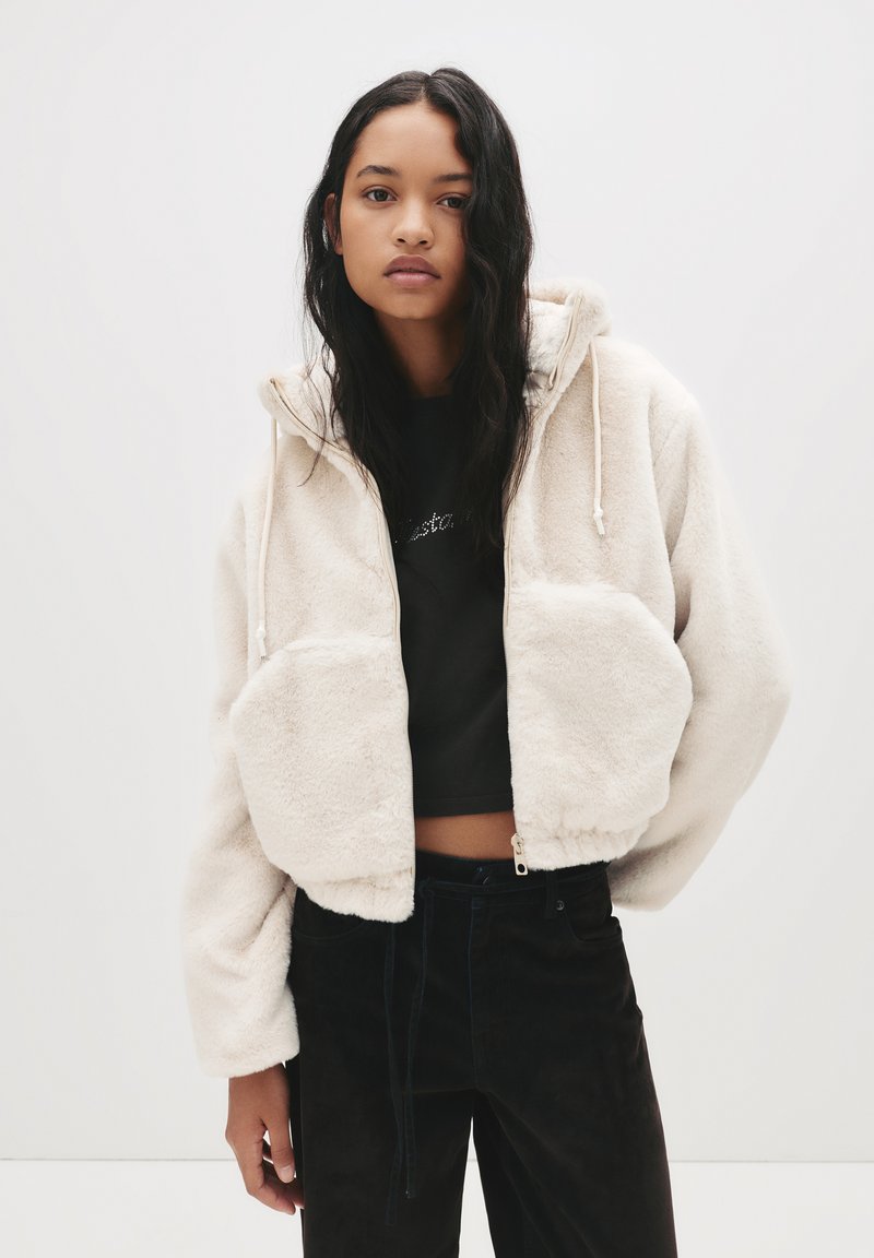 Mlada ženska z dolgimi temnimi lasmi, oblečena v kremno puhasto zadrgo s kapuco čez črn kratki top in črne hlače, stoji pred enobarvno belo ozadjem.