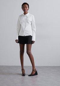 White long-sleeve shirt with button closure and pleated black mini skirt; model wears black patent wedge shoes with ankle straps.