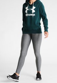 Osoba oblečená v tmavě zelené mikině Under Armour, šedých legínách a černých sportovních botách, stojící s jednou nohou vystrčenou dopředu.