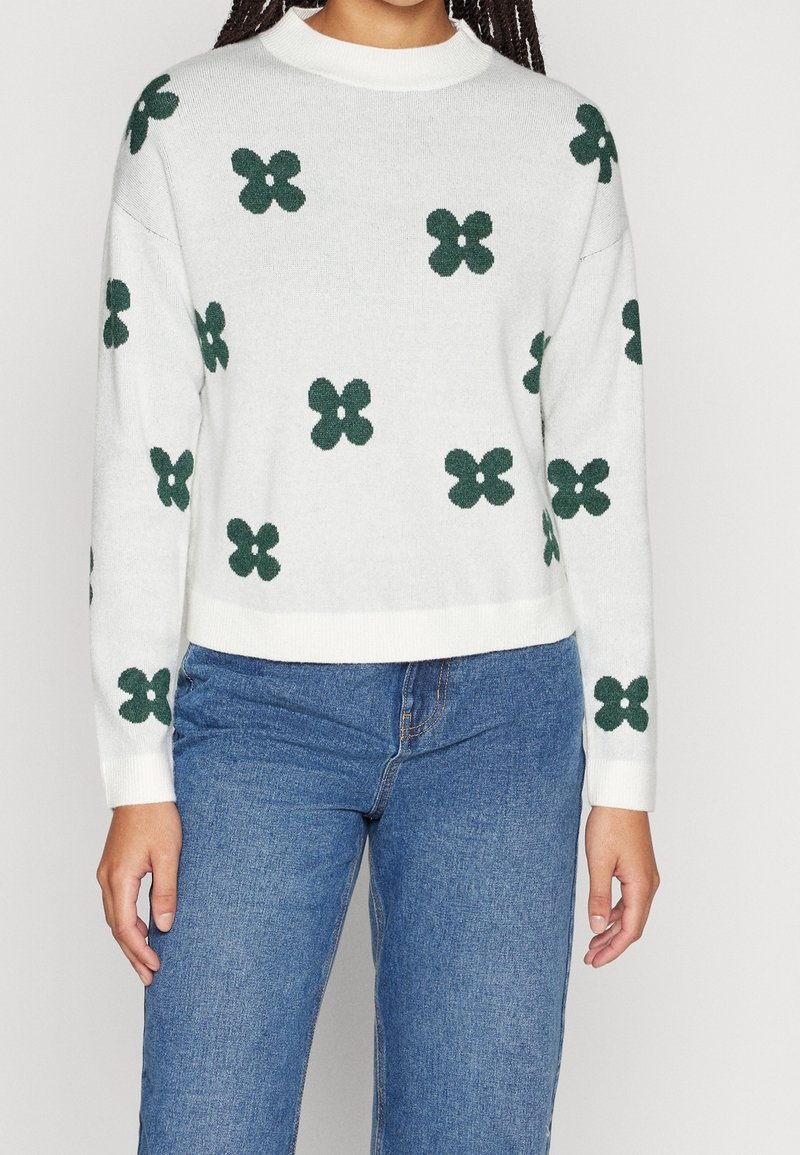 Personne portant un pull crème avec des motifs floraux à quatre pétales vert foncé, associé à un jean en denim bleu, visible du cou aux hanches.