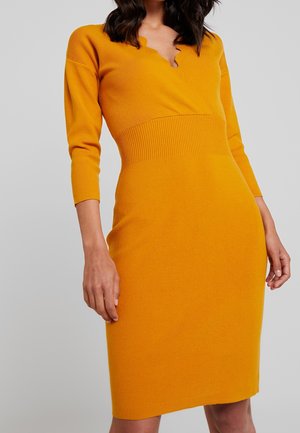 Mujer con un vestido de punto amarillo mostaza ajustado, con mangas tres cuartos y escote en V con borde en ondas, sobre un fondo liso.