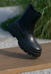 Černá kotníková bota vyrobená z lesklé kůže s tlustou, texturovanou podrážkou, elastickými bočními panely a kulatým špičatým designem, vystavená na dřevěném povrchu.