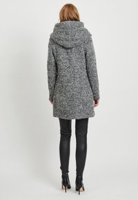 Donna che indossa un cappotto grigio strutturato di lunghezza media con cappuccio, pantaloni in pelle nera e stivaletti neri con tacco alto, in piedi di spalle.