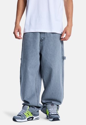 Osoba nosząca luźne, jasnoniebieskie jeansy, szare i neonowo zielone trampki oraz białą koszulę, stojąca na jednolitym tle.