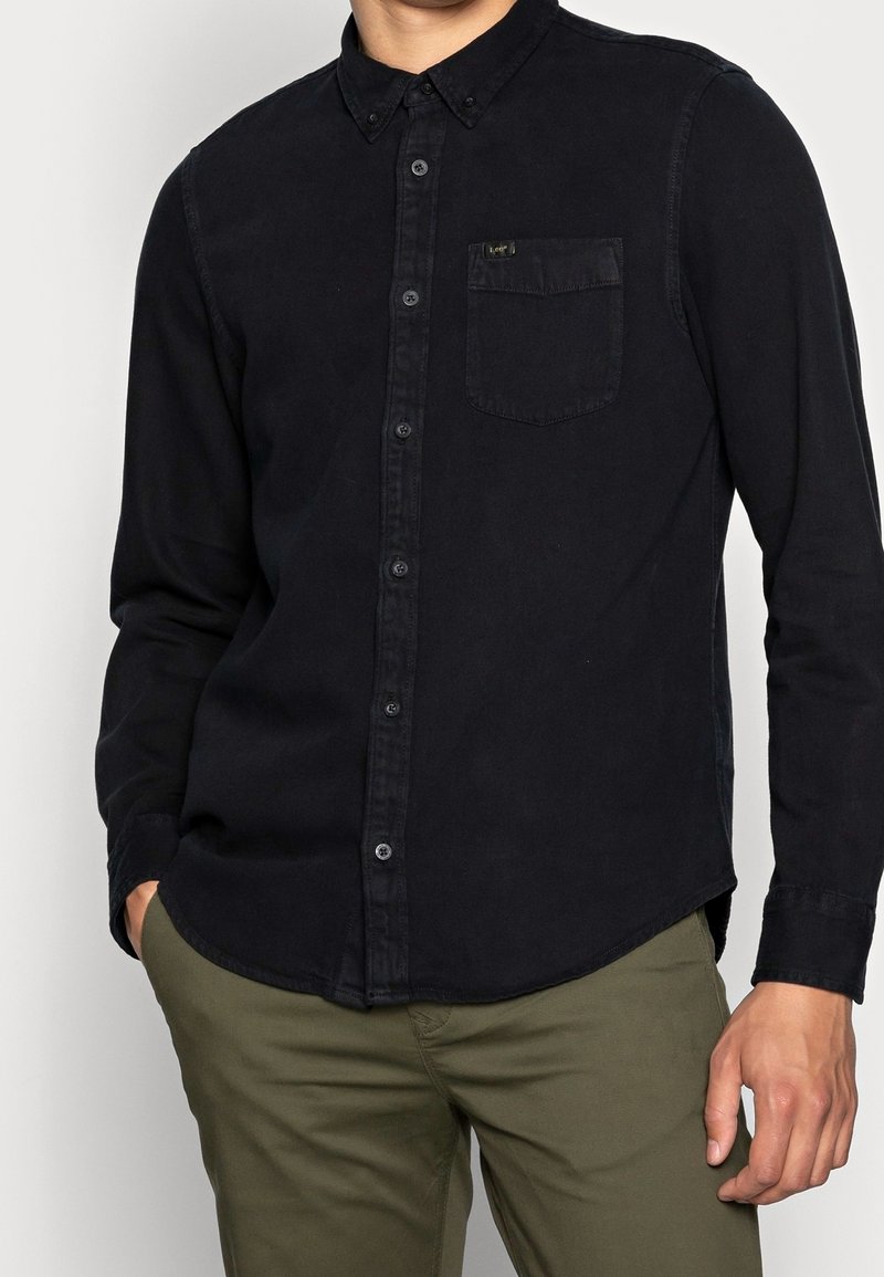 Homme portant une chemise noire à manches longues boutonnée avec une poche poitrine et un pantalon vert olive, les mains partiellement dans les poches.