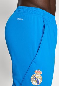 Blå sportsshorts med snor i taljen, sidelomme med lynlås og Real Madrid-logo med krone mønster. Fremstillet af letvægtsstof.