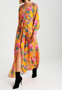 Maxiklänning i ljusgul med rosa blommönster och gröna löv, djup V-ringning, bälte i midjan, långa ärmar och sidoklaff.