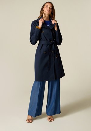 Femme portant un trench-coat bleu marine foncé ceinturé par-dessus un jean bleu à jambes larges et des sandales ouvertes sur un fond clair uni.