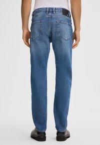 Jeans in denim blu con una vestibilità rilassata, cuciture visibili, cinque tasche e una patch nera con logo nella parte posteriore della vita. I pantaloni presentano un lavaggio sbiadito.