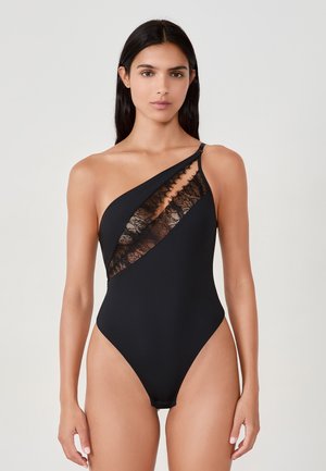 Femme portant un maillot de bain noir à une épaule avec un panneau en dentelle diagonale sur la poitrine, se tenant contre un fond clair.