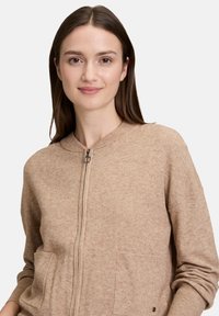 Junge Frau mit glattem braunem Haar, die einen beigen Reißverschluss-Pullover mit Taschen trägt und sanft vor einem schlichten hellen Hintergrund lächelt.