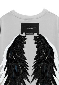 Camiseta gris de manga corta con diseños de alas en blanco y negro hechos de una tela con textura brillante; etiqueta adjunta en el escote.