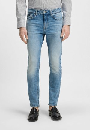 Mężczyzna w jasnoniebieskich obcisłych dżinsach, szarej koszuli z guzikami oraz czarnych skórzanych mokasynach, stojący na jednolitym tle.