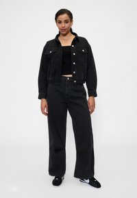 Čierna rifľová bunda s gombíkmi na zapínanie, dvoma náprsnými vreckami a elastickým lemom, v kombinácii s čiernymi širokými džínsami s vysokým pásom a čiernymi teniskami.