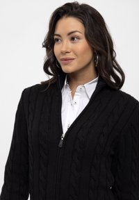 Sort kabelstrikket sweater med lynlås foran og høj krave, med et tekstureret mønster. Bæres med en hvid skjorte med krave under.