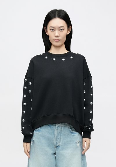 Sweatshirt noir avec des clous argentés sur l'encolure et le long des manches, coupe décontractée et poignets côtelés. Porté avec un jean bleu clair.
