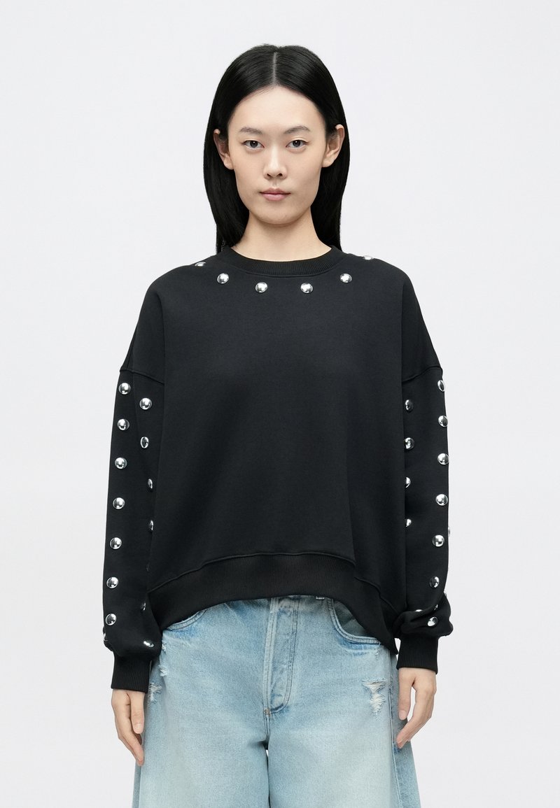 Čierna mikina s striebornými cvokmi na výstrihu a pozdĺž rukávov, voľného strihu s rebrovanými manžetami. Nosená so svetlomodrými džínsami.