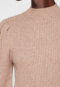 Gros plan d'une personne portant un pull beige en maille côtelée avec un col montant et un léger détail bouffant à l'épaule.