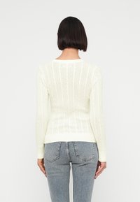 Femme aux cheveux courts et foncés portant un pull en maille torsadée crème et un jean bleu clair, vue de dos sur un fond uni.