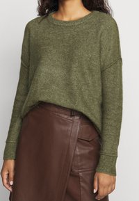 Pull en maille vert avec un large col rond et des poignets côtelés, associé à une jupe en cuir marron présentant un design asymétrique et un détail de poche.