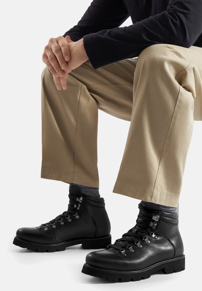 Czarne skórzane buty turystyczne z metalowymi oczkami, sznurowadłami i fakturowaną powierzchnią. Noszone z szarymi skarpetami i beżowymi spodniami. Prosty design, solidna podeszwa.