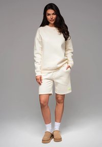 Completo di felpa e pantaloni corti color crema, con polsini a coste e tasche laterali, abbinato a ciabatte beige e calze bianche.