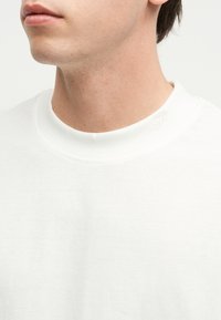 Κοντινό πλάνο του κάτω μέρους του προσώπου και του λαιμού ενός ατόμου που φοράει μια απλή λευκή μπλούζα με στρογγυλή λαιμόκοψη και μια μικρή, διακριτική κεντημένη αρχική κοντά στο γιακά.