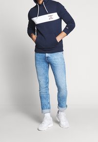 Mężczyzna w granatowym hoodie z napisem "Tommy Jeans", jasnoniebieskich podwiniętych dżinsach, białych skarpetach i białych sneakersach, stojący z rękami w kieszeniach.