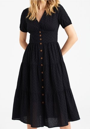 Frau trägt ein schwarzes Midikleid mit Lochmuster, kurzen Ärmeln, V-Ausschnitt, Knopfleiste vorne und stufigem Rock vor schlichter weißer Hintergrund.