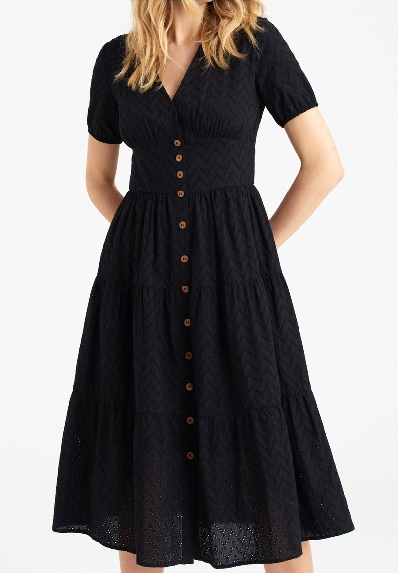 Femme portant une robe midi noire à œillets, manches courtes, col en V, boutonnée devant, et jupe à volants, sur fond blanc neutre.