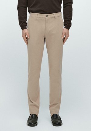 Mężczyzna w beżowych spodniach o prostych nogawkach, czarnych mokasynach i ciemnobrązowej bluzce z długim rękawem, stojący na jednolitym tle.