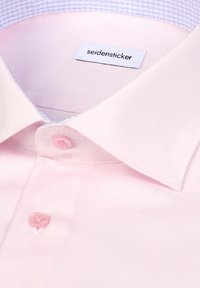 Chemise rose pâle en tissu texturé, avec un col pointu, deux boutons et un col intérieur à carreaux bleus. Étiquette de la marque visible.