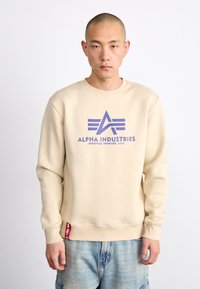 Beige kapucnis pulóver lila Alpha Industries logóval. Rászorított mandzsetták és szegély, oldalsó címke részlet, és laza fazon. Puhán sima pamut keverék anyag.