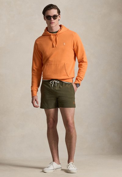 Άνδρας με πορτοκαλί φούτερ με κουκούλα, χακί σορτς, λευκά αθλητικά παπούτσια, γυαλιά ηλίου και ρολόι, στέκεται μπροστά σε ουδέτερο φόντο.