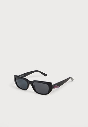 Lunettes de soleil rectangulaires noires avec verres foncés et logo coloré "Guess" sur la branche contre un fond blanc.