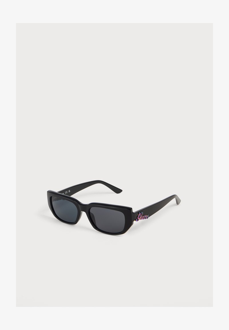Lunettes de soleil rectangulaires noires avec verres foncés et logo coloré "Guess" sur la branche contre un fond blanc.