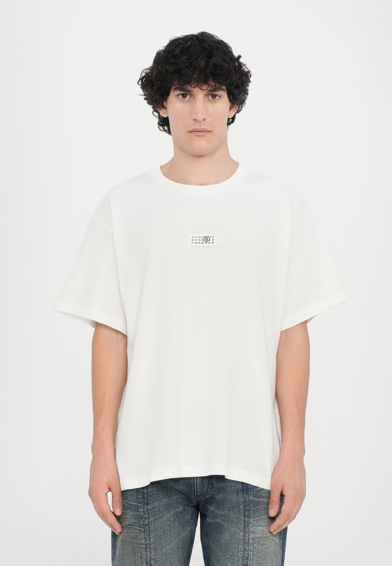 T-shirt en coton blanc oversize à col rond et manches courtes, avec une petite impression graphique sur la poitrine gauche.