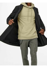 Charcoal-Wollmantel offen über einem cremefarbenen Hoodie mit Kängurutasche. Sichtbare graue Karohose, die einen schlanken, modernen Lagenlook zeigt.